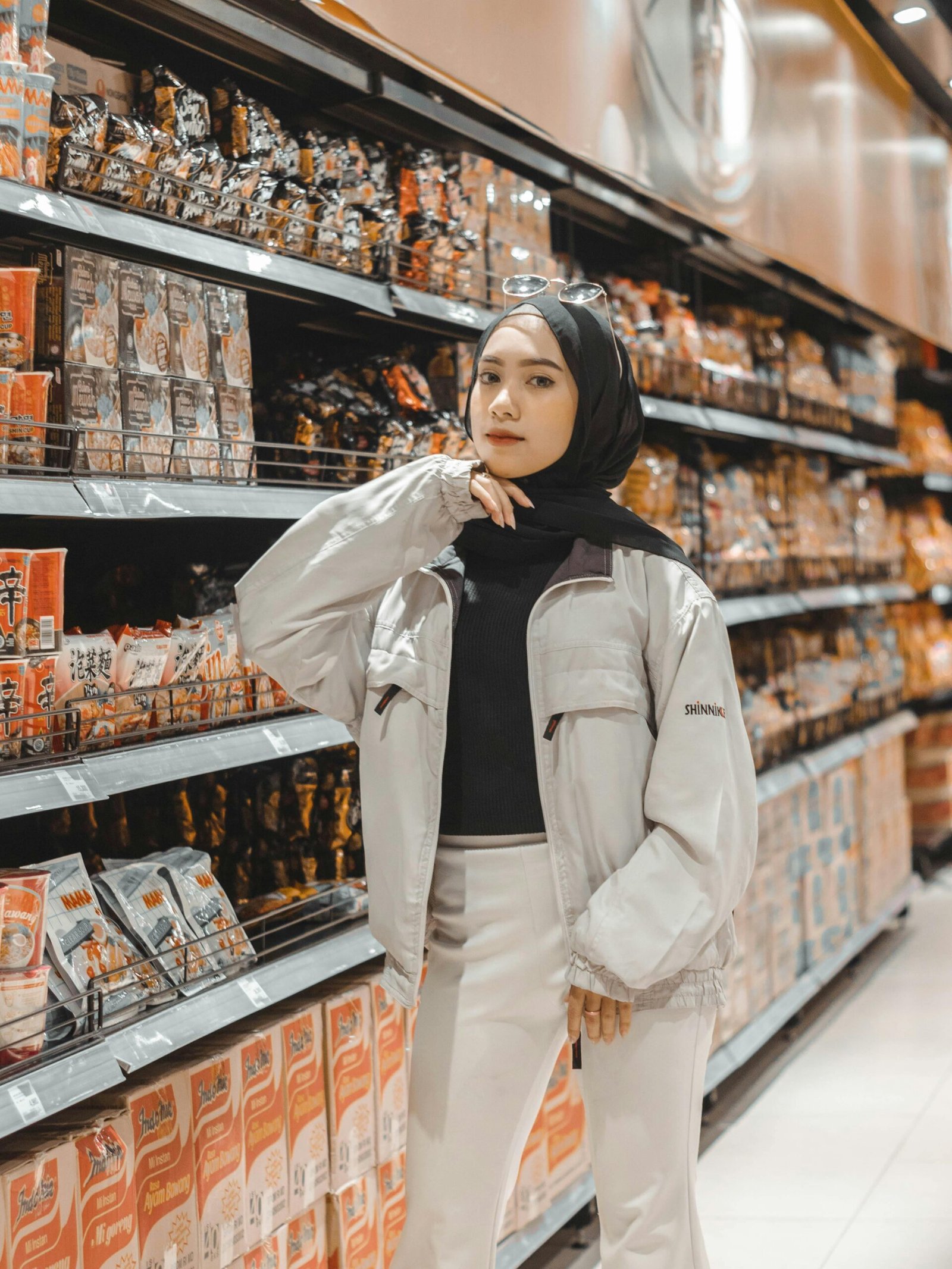 Confident young Muslim lady in stylish outfit and hijab standing near shelves in supermarket and choosing various products